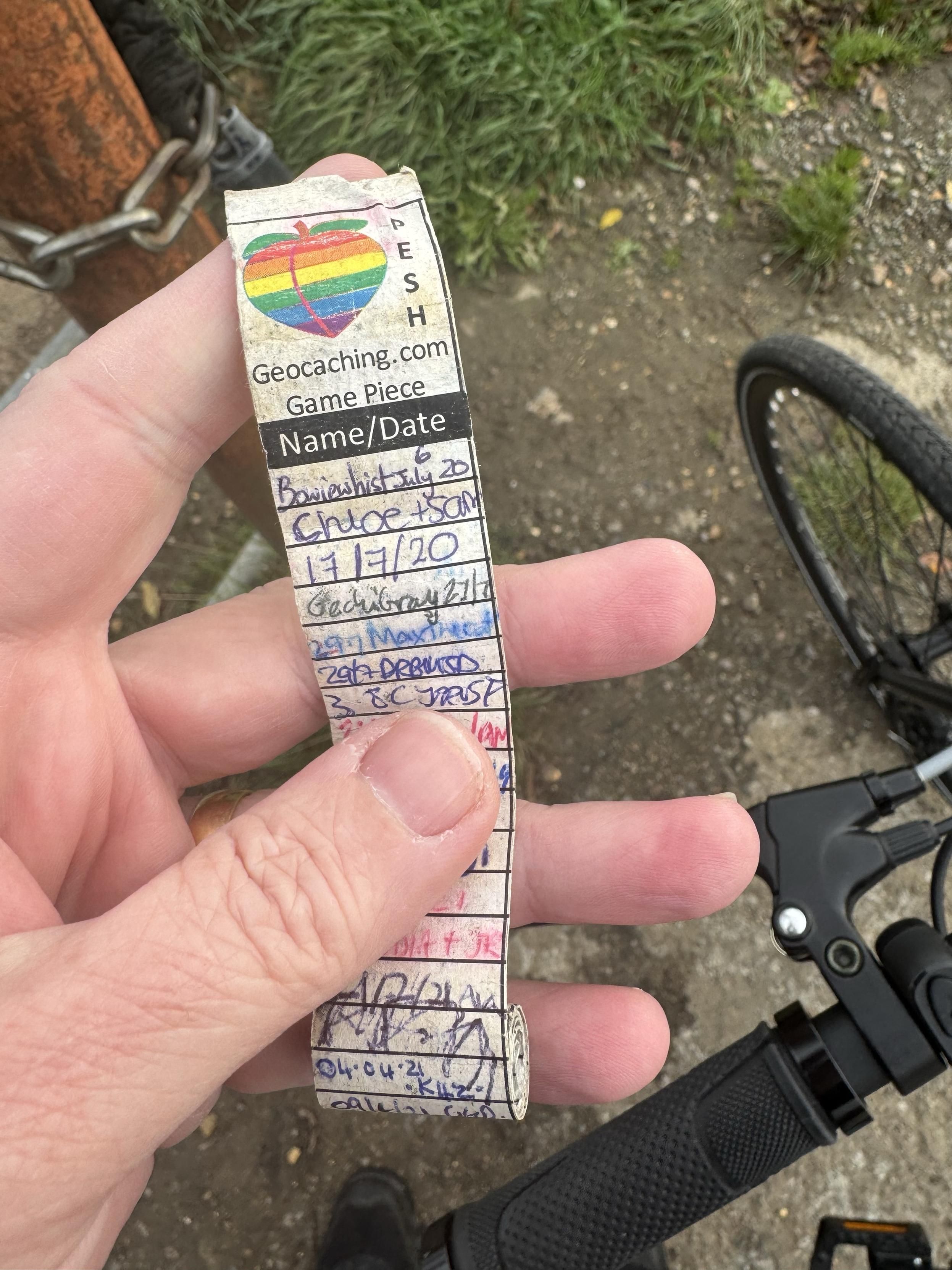 A hand holds a geocaching logbook with various names and dates written on it, alongside a rainbow heart logo. The background features grass and part of a bicycle.