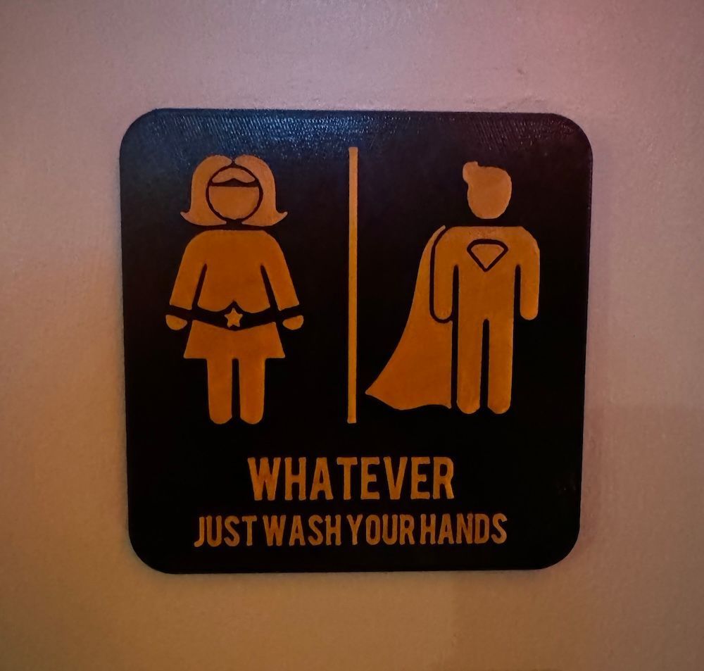 A toilet door sign showing female and mail superheroes. It reads 